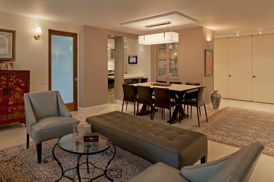 Open living and dining area with gray armchairs, a tufted bench, a dining table under a rectangular pendant and a glass-front cabinet